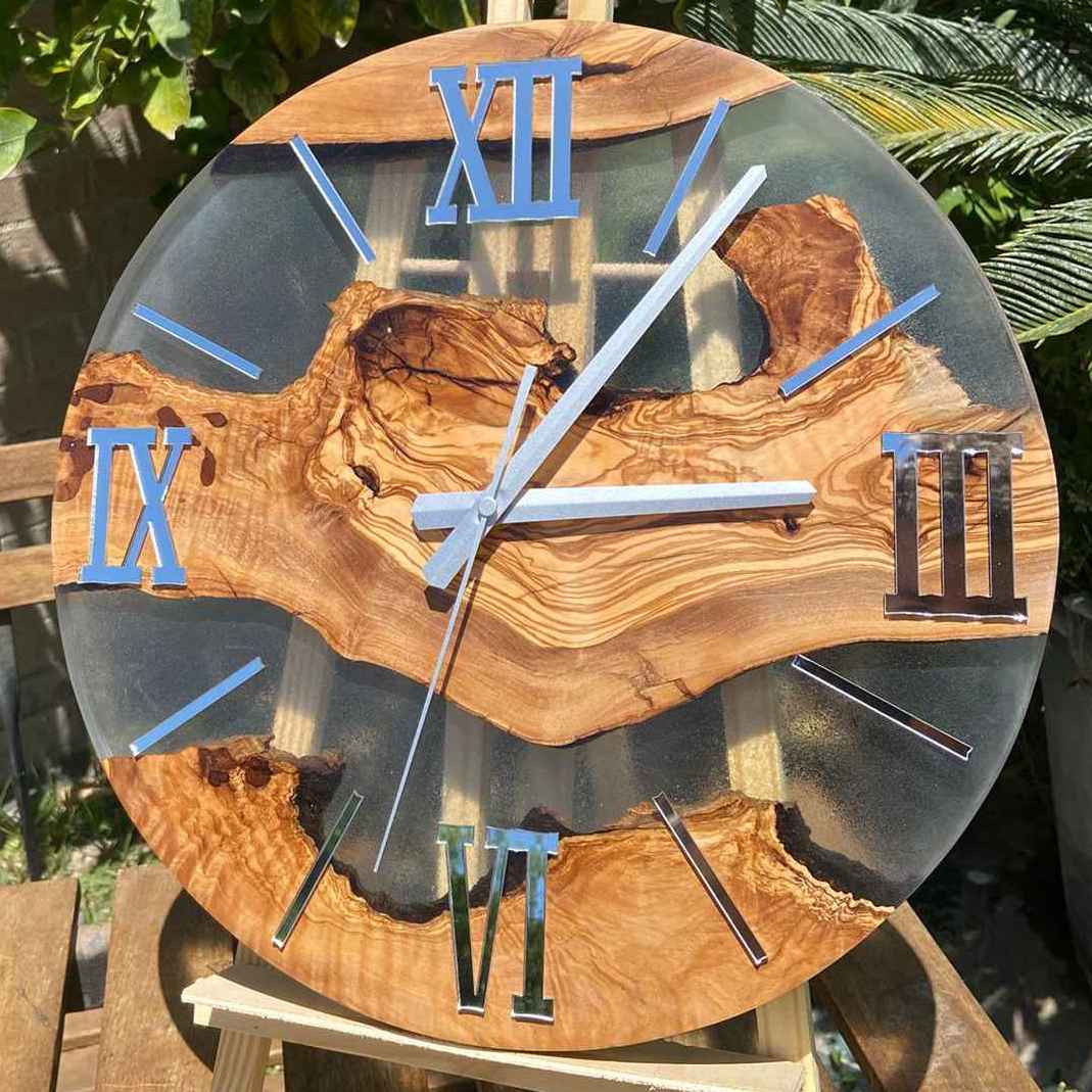 Primo piano ravvicinato di un orologio da parete artigianale in legno di ulivo e resina trasparente. L'illuminazione naturale all'aperto fa risaltare le venature calde del legno e i riflessi specchiati delle lancette e dei numeri romani argentati.