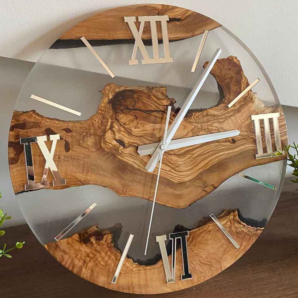 Orologio da parete artigianale realizzato con una tavola di pregiato legno di ulivo immersa in resina trasparente, impreziosito da lancette e numeri romani con finitura argentata a specchio.