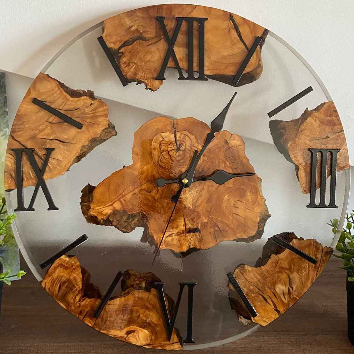 Orologio da parete artigianale realizzato in radica di ulivo e resina trasparente, con eleganti numeri romani neri. L'orologio è fotografato frontalmente, appoggiato su un mobile in legno tra due piccole piantine. L'orologio va appeso al muro ed è indicato per arredamenti chiari, è stato realizzato a roma da noi e personalizzato in base alle richieste del cliente. Noi siamo MM Wood and resin