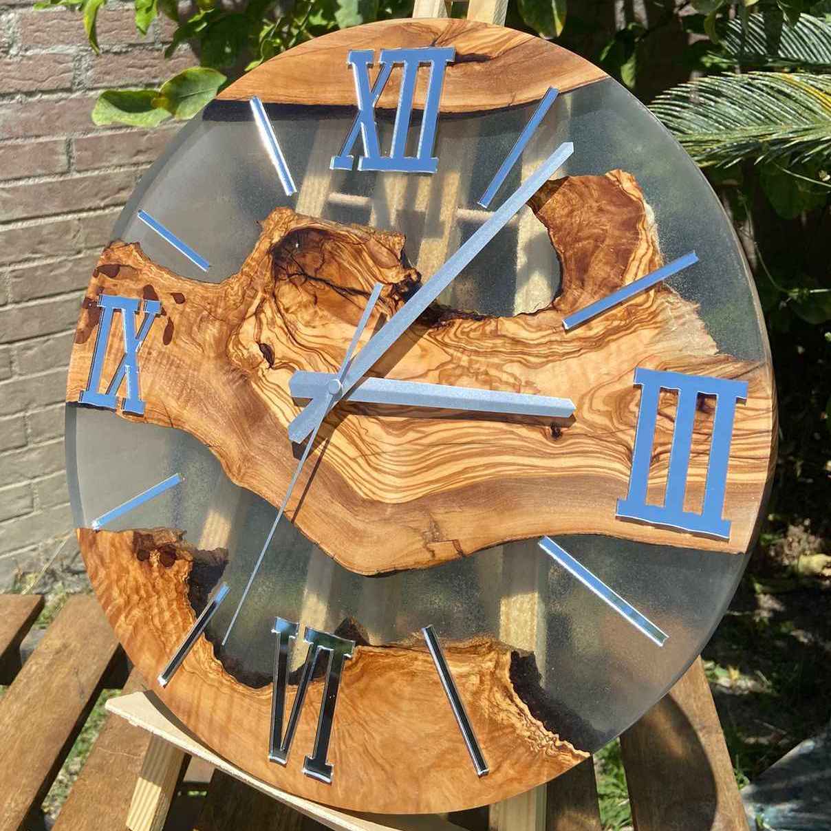 Orologio da parete artigianale in pregiato legno di ulivo e resina trasparente, con numeri romani specchiati argento. L'orologio è esposto all'aperto su un piccolo cavalletto di legno, illuminato dalla luce naturale del sole con piante verdi sullo sfondo.