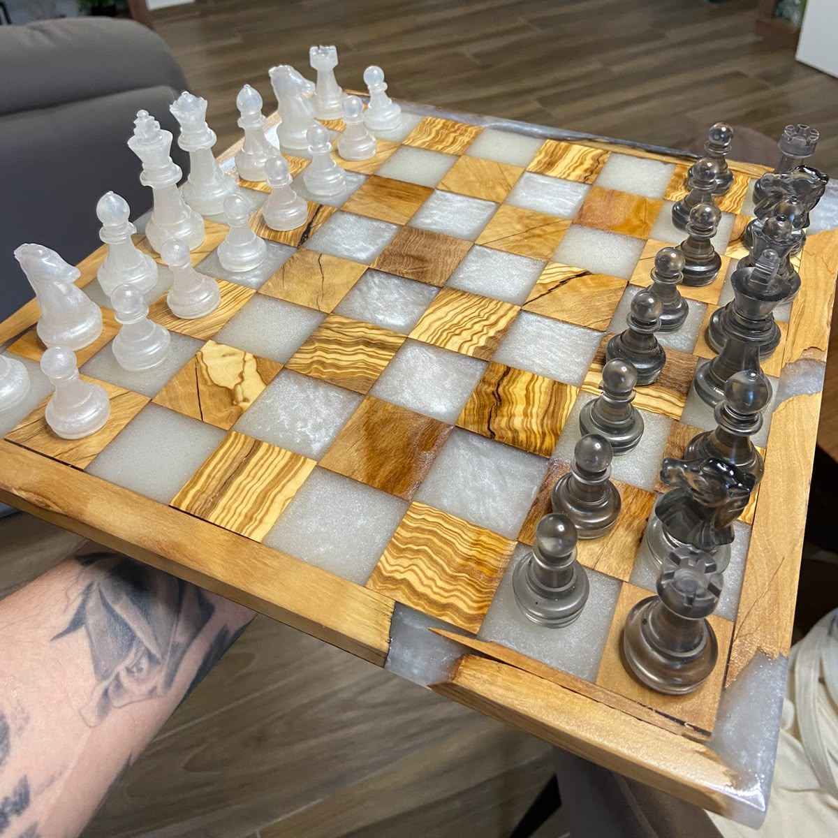Scacchiera quadrata in legno di ulivo e resina bianca tenuta in mano, con pezzi bianchi e neri posizionati sulla tavola. Il produttore siamo noi, Marco Morrone wood and resin, unici nel nostro lavoro. Creiamo pezzi personalizzati a Roma e spediamo in tutta italia.