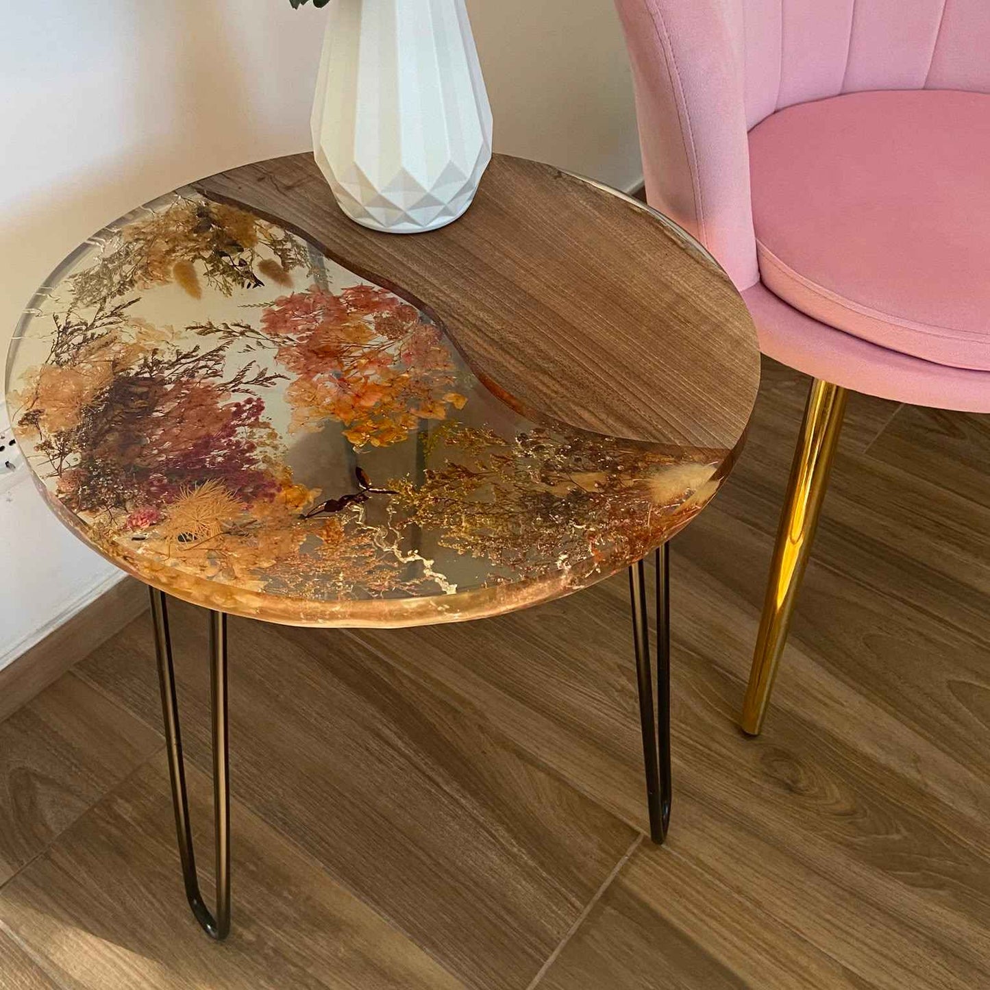 Tavolino da salotto rotondo in stile industriale e botanico, realizzato in legno di noce e resina trasparente con fiori secchi incastonati. Il tavolo ha gambe in metallo ed è affiancato a una poltroncina rosa su un pavimento in gress