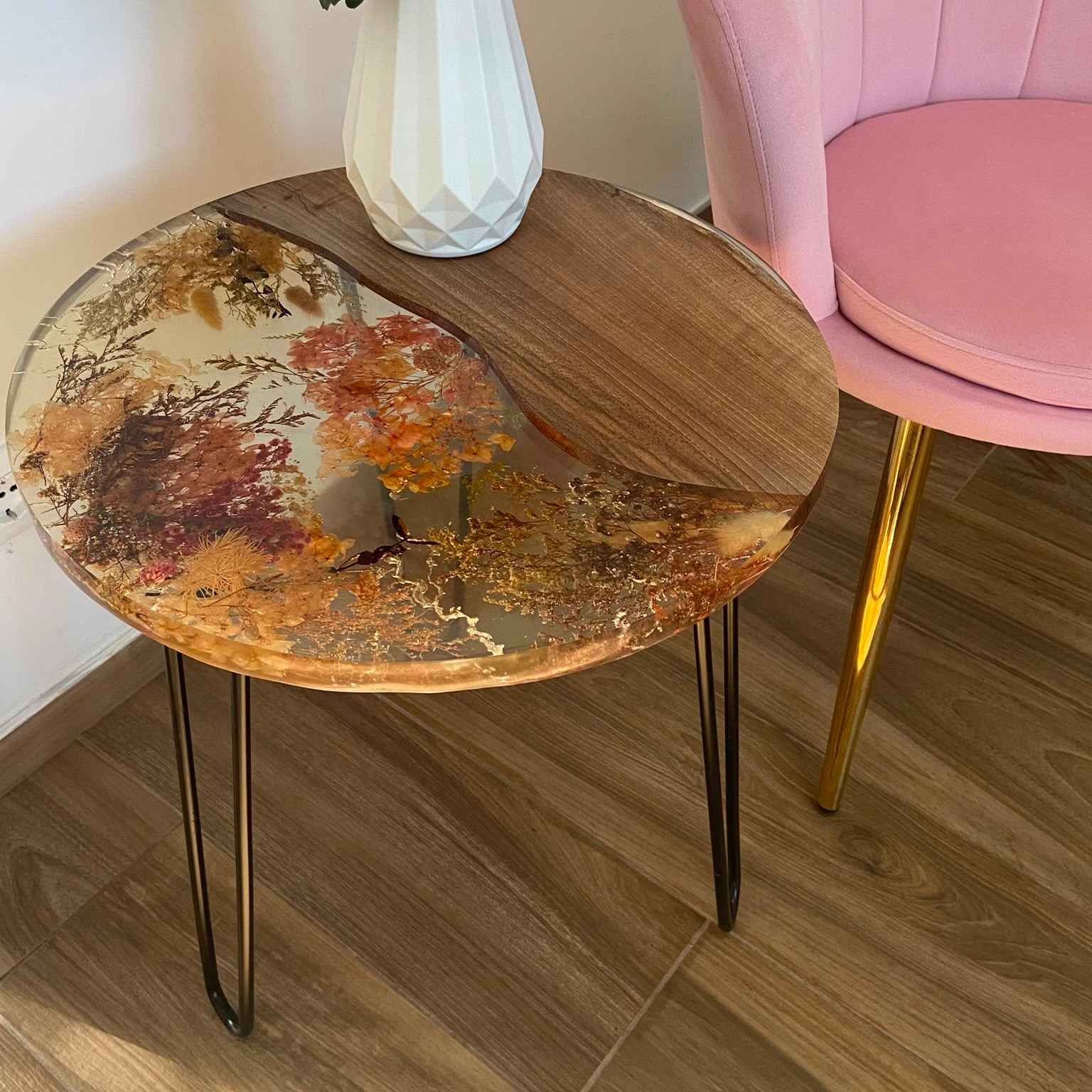 Tavolino da salotto rotondo in stile industriale e botanico, realizzato in legno di noce e resina trasparente con fiori secchi incastonati. Il tavolo ha gambe in metallo ed è affiancato a una poltroncina rosa su un pavimento in gress