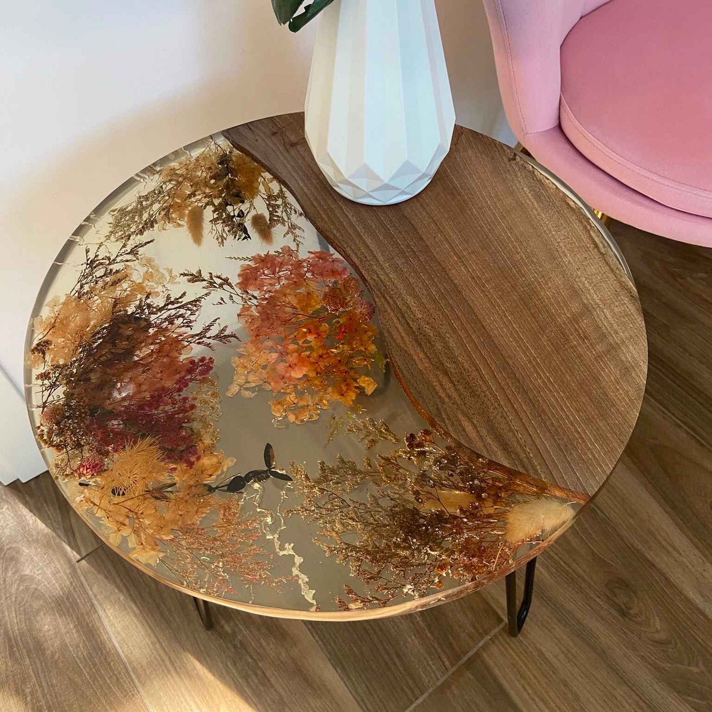 Tavolino da caffè rotondo artigianale in legno di noce e resina epossidica trasparente con fiori secchi incorporati, posizionato su un pavimento in legno.