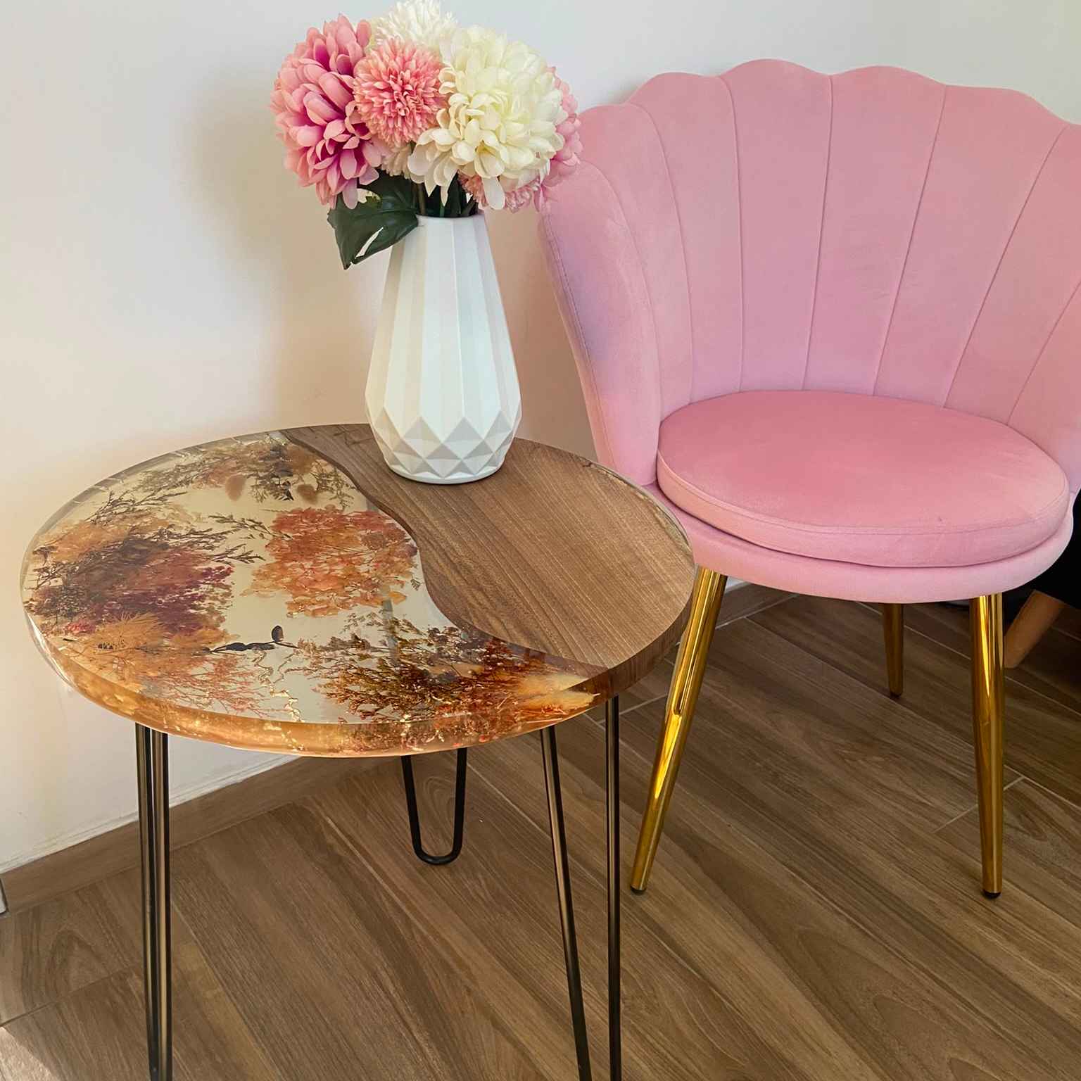 Tavolino da salotto rotondo in legno e resina trasparente con fiori secchi incorporati. Sul piano poggia un vaso geometrico bianco con fiori, accanto a una poltroncina a conchiglia in velluto rosa.