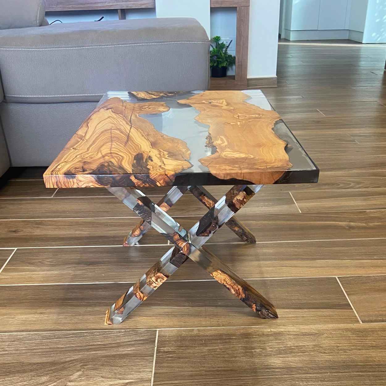 Vista laterale di un tavolino da salotto in legno di ulivo e resina trasparente creato da noi a roma, MM Wood and resin, caratterizzato da gambe incrociate a X, posizionato accanto a un divano grigio.