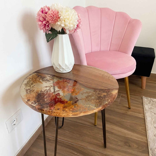 Tavolino da salotto rotondo artigianale in legno di noce e resina trasparente con fiori secchi incorporati. Sul tavolo poggia un vaso bianco con fiori, accanto a una poltroncina in velluto rosa e un pouf nero.