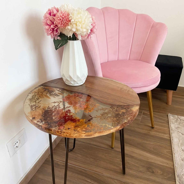 Tavolino da salotto rotondo artigianale in legno di noce e resina trasparente con fiori secchi incorporati. Sul tavolo poggia un vaso bianco con fiori, accanto a una poltroncina in velluto rosa e un pouf nero.