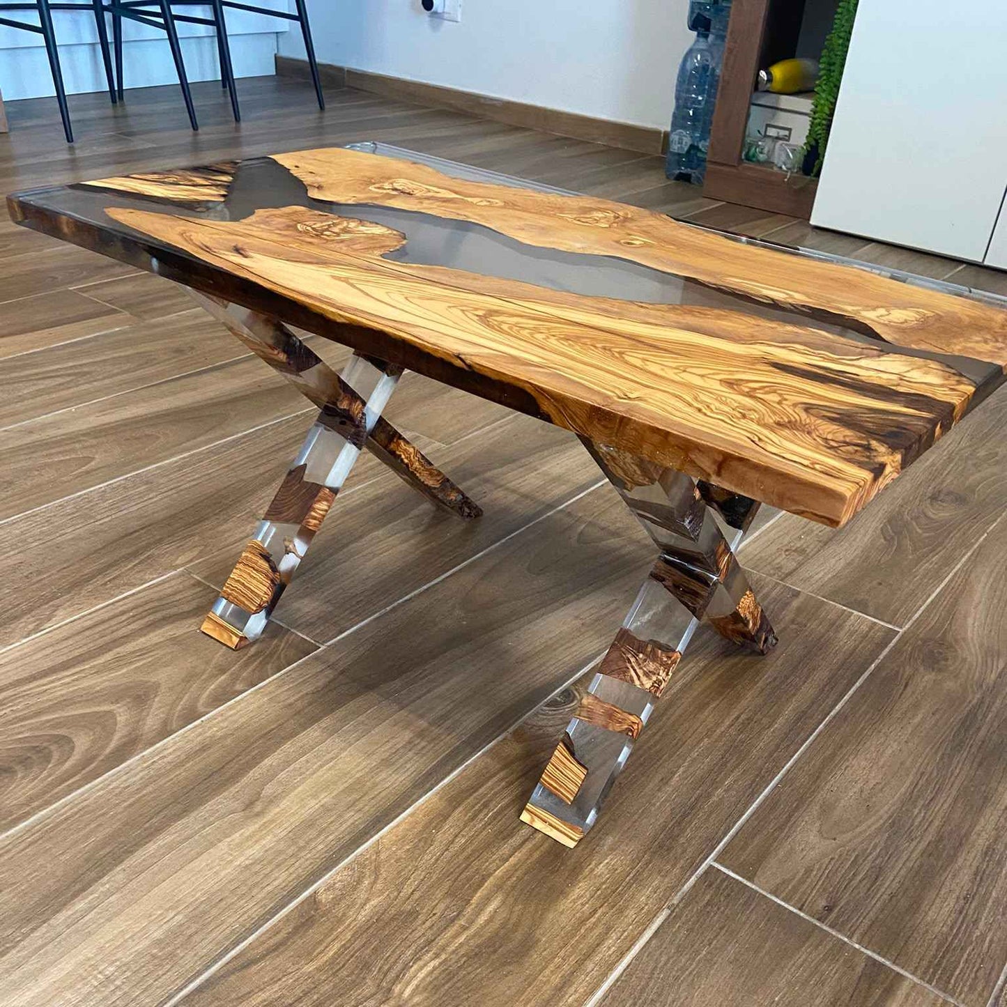 Tavolino da salotto rettangolare in stile river table, realizzato artigianalmente con legno di ulivo e resina trasparente. Il tavolo si distingue per le originali gambe a incrocio (X), anch'esse composte da un mix di legno e resina, posizionato su un pavimento in gres porcellanato.