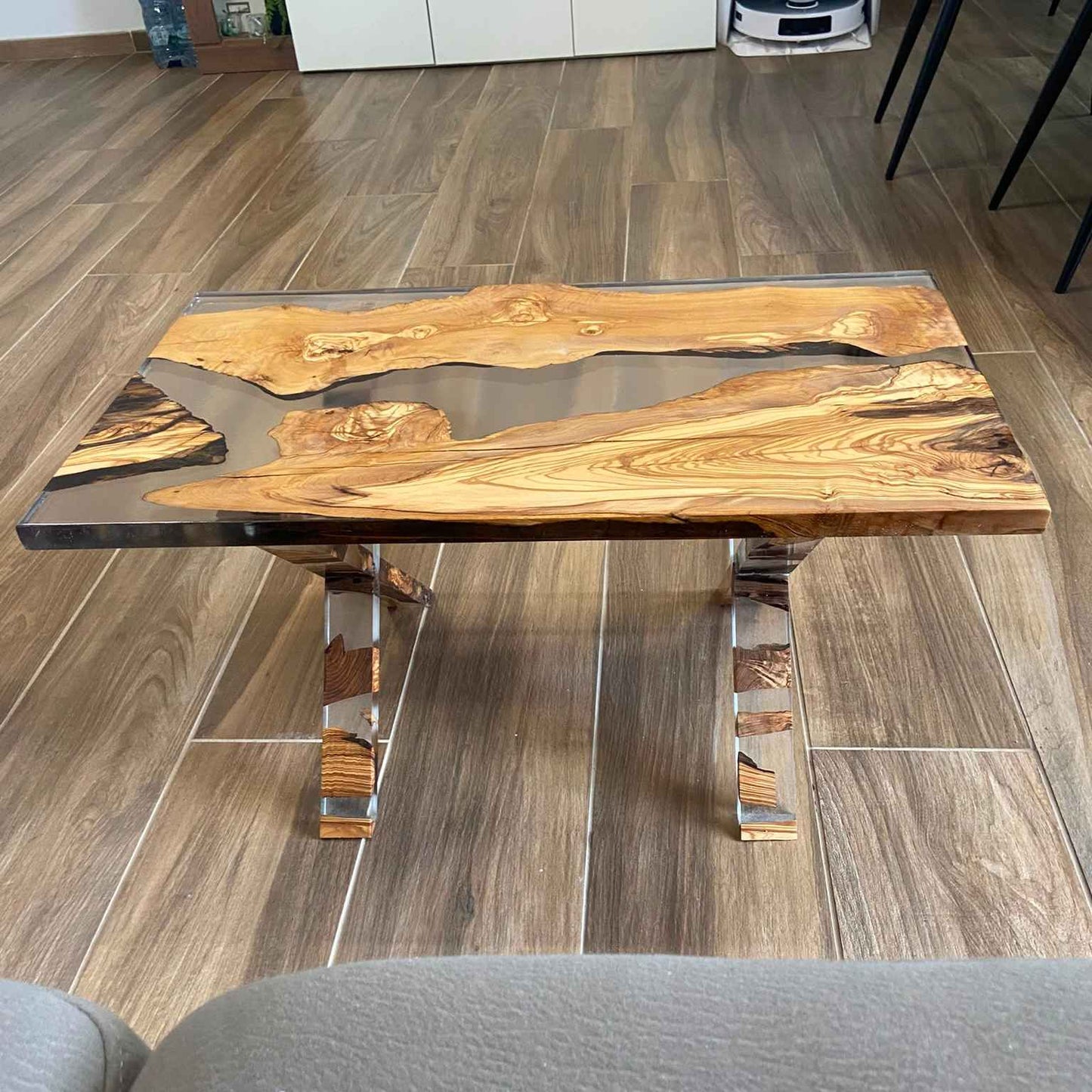 Vista frontale e dall'alto di un tavolino artigianale da salotto in stile river table, realizzato con tavole di legno di ulivo e resina trasparente, che lascia intravedere le gambe incrociate sottostanti.